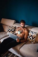 Mid adult man with tattooed chest on sofa with lap